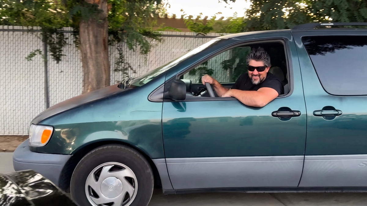 A smiling driver leaning out of the window of a teal minivan.